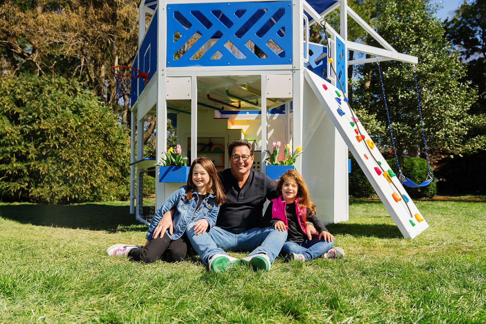 Photo of a man and two children sitting in the grass smiling in front of the Blue Squirrel playground.